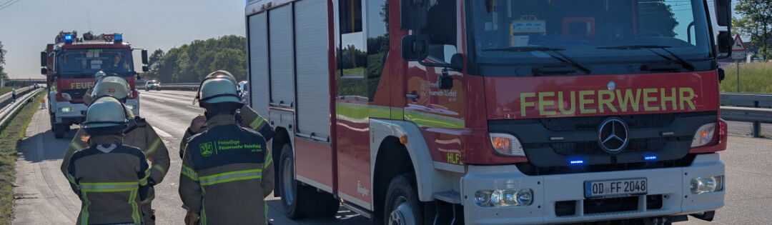 Ein Feuerwehrfahrzeug der FF Reinfeld steht mit eingeschaltetem Blaulicht auf einer asphaltierten Straße. Im Vordergrund stehen mehrere Feuerwehrleute in Schutzkleidung mit dem Rücken zur Kamera. Im Hintergrund ist ein weiteres Feuerwehrfahrzeug zu sehen.